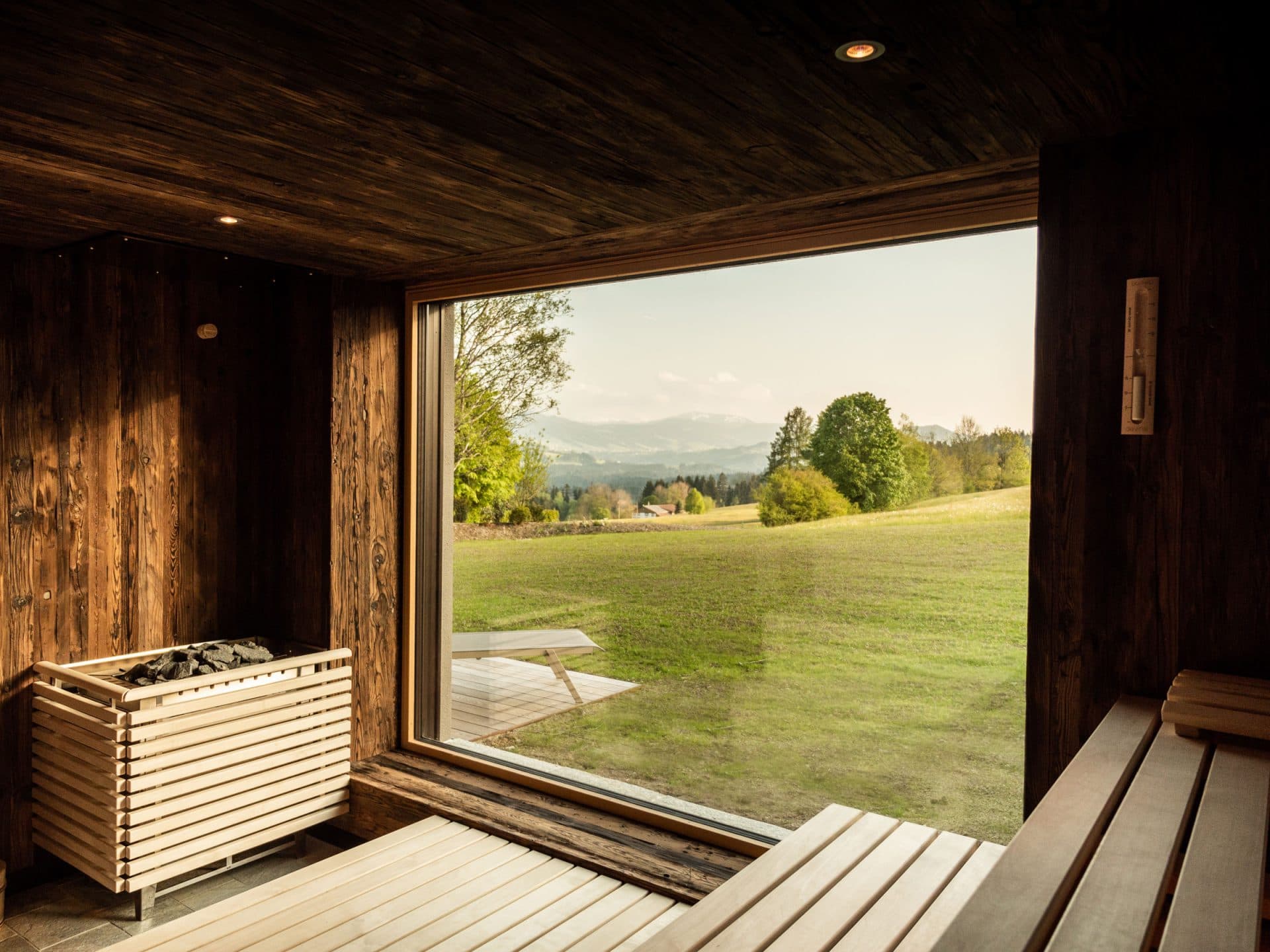 Panorama-Sauna in dunklem Altholz, Saunaofen mit Steinen, bodentiefes Fenster zur Weide und den Bergen dahinter