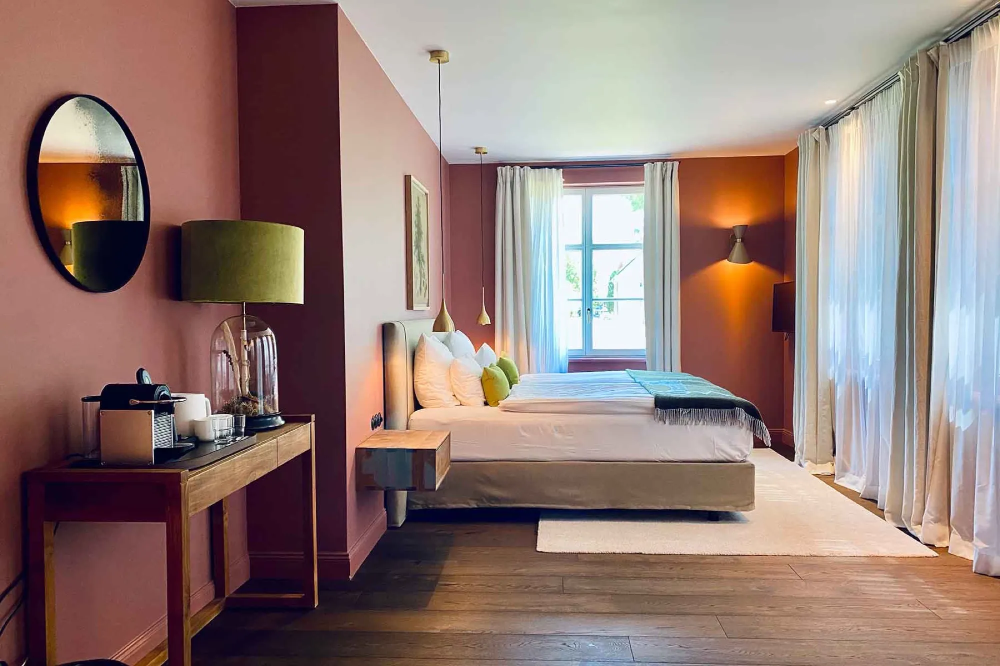 Übersicht der Suite Eins mit Bett vor rosafarbener Wand, Konsolentisch mit Rundspiegel, hohe Fenster mit Vorhängen
